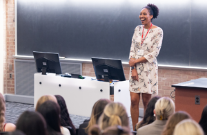 Professional woman in a floral dress with a red lanyard presenting to students in a lecture hall, standing beside two monitors at a podium.