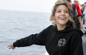 Smiling young woman in a black hoodie with arms outstretched on a boat, the ocean in the background.