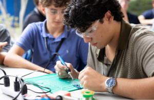 Two young men soldering electronic components on a green prototyping board outdoors, wearing safety goggles.