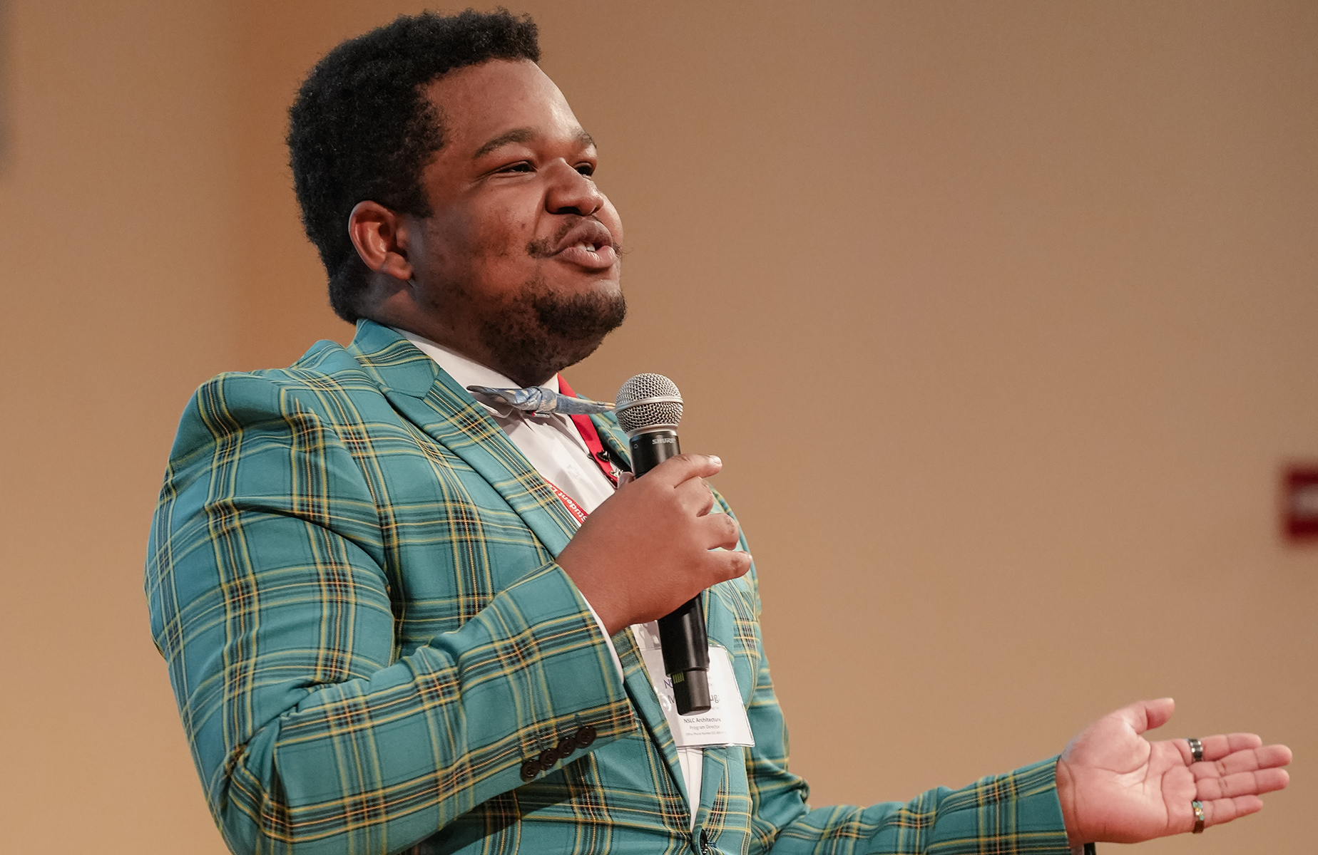 Man in teal plaid suit speaks into a handheld microphone on stage, gesturing with his left hand, wearing a name badge.