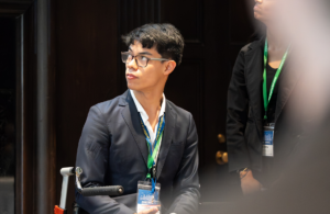 Man in a dark blazer and glasses looking to the left at a conference, wearing a blue-green lanyard with a badge.