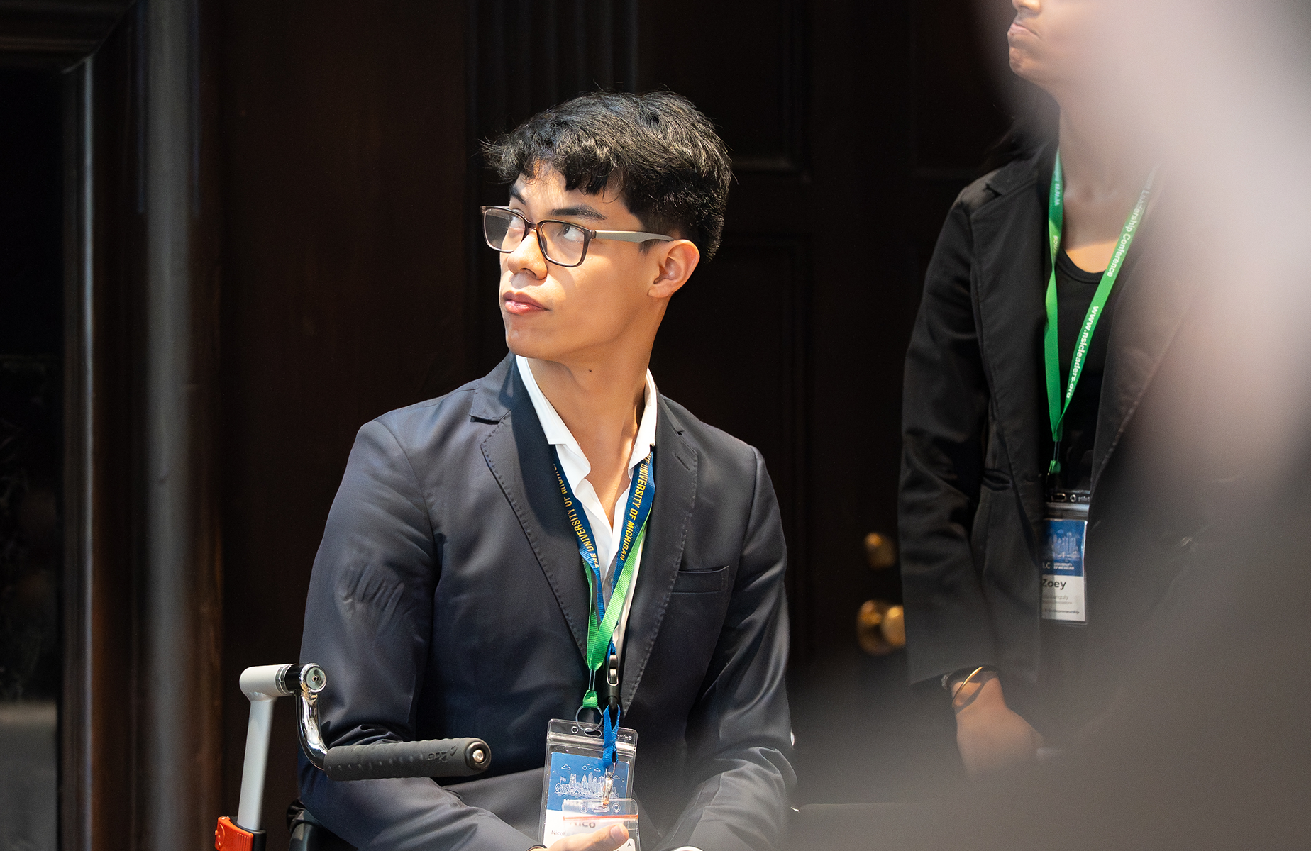 Man in a dark blazer and glasses looking to the left at a conference, wearing a blue-green lanyard with a badge.