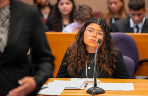 Young woman with glasses speaking into a microphone at a conference table, papers in the foreground.