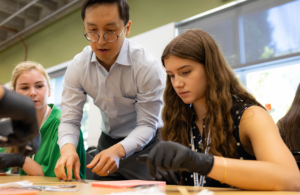 A man with glasses and a woman with long hair lean over a table, examining materials during a workshop.